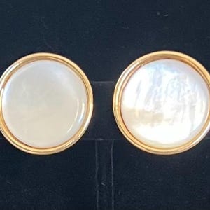 May include: A pair of circular earrings featuring a smooth, iridescent white mother-of-pearl inlay set within a polished gold-tone metal frame. The design is simple and round, highlighting the natural luster of the shell material.