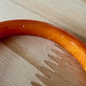May include: A smooth, semi-translucent orange bangle bracelet with a rounded profile. The material exhibits subtle variations in color density and internal marbling, creating a warm, glowing appearance across the curved surface of the circular jewelry piece.