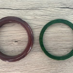 May include: Two circular bangle bracelets. One bracelet is a deep, dark brown with a textured, scale-like pattern across its surface. The second bracelet is a solid, translucent dark green with a smooth, polished finish and subtle color variations.