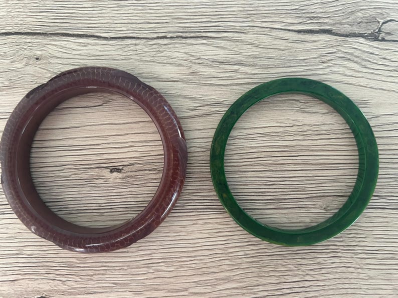 May include: Two circular bangle bracelets. One bracelet is a deep, dark brown with a textured, scale-like pattern across its surface. The second bracelet is a solid, translucent dark green with a smooth, polished finish and subtle color variations.