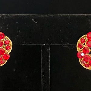 May include: A pair of round gold-tone earrings featuring a cluster of vibrant red faceted crystals. Each earring has a large central red stone surrounded by smaller red stones set in a circular gold-tone metal frame with a textured finish.