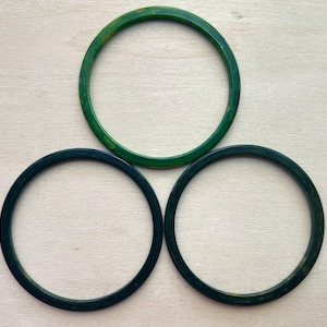 May include: Three circular bangle bracelets arranged in a triangular formation. One bracelet is a bright, translucent emerald green, while the other two are a darker, solid forest green. Each bangle features a smooth, polished, rounded surface.