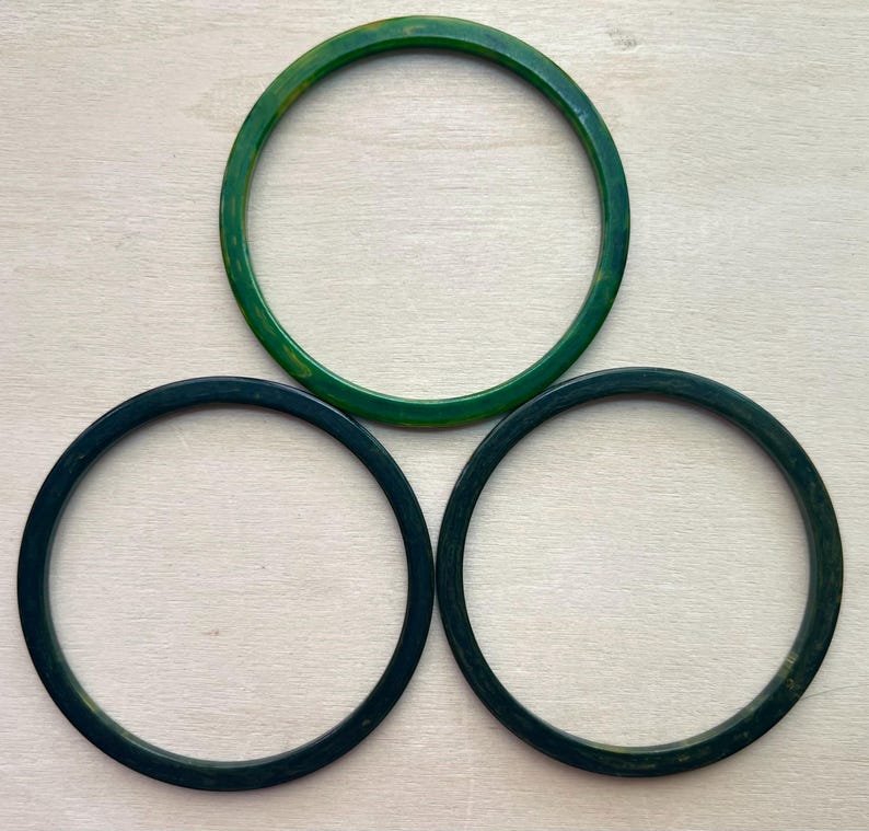 May include: Three circular bangle bracelets arranged in a triangular formation. One bracelet is a bright, translucent emerald green, while the other two are a darker, solid forest green. Each bangle features a smooth, polished, rounded surface.