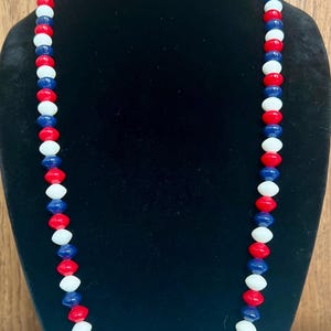 May include: A long beaded necklace with alternating red, white, and blue beads. The necklace is displayed on a black mannequin, showcasing the patriotic color scheme. The beads are oval-shaped.