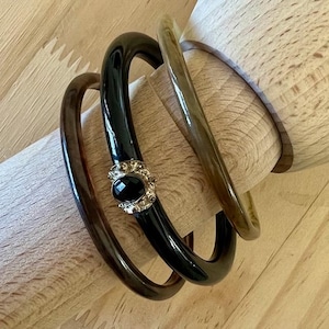 May include: Three smooth, rounded bangles stacked together. One is dark brown, one is a translucent light tan, and the central black bangle features a silver-toned metal accent piece set with a single, polished, oval-shaped black stone.