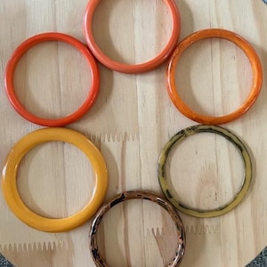 May include: A collection of six circular bangle bracelets arranged in a cluster. The set includes two solid orange bangles, one solid yellow bangle, one translucent orange bangle, one beige bangle with dark brown marbling, and one brown patterned bangle.