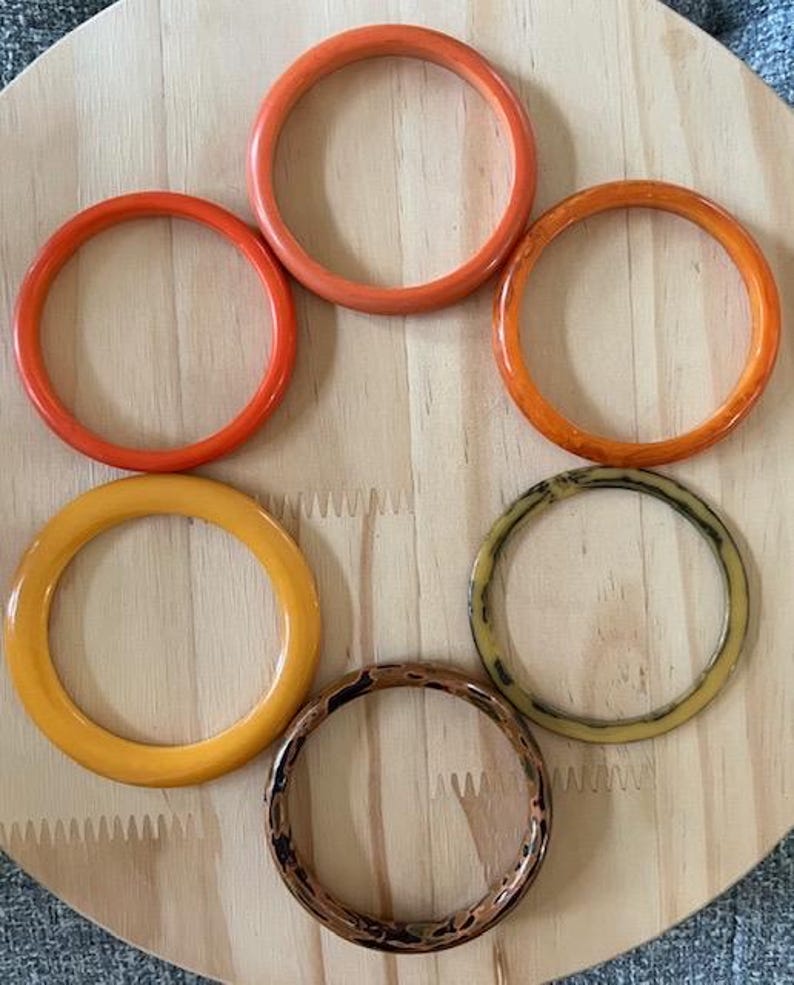 May include: A collection of six circular bangle bracelets arranged in a cluster. The set includes two solid orange bangles, one solid yellow bangle, one translucent orange bangle, one beige bangle with dark brown marbling, and one brown patterned bangle.