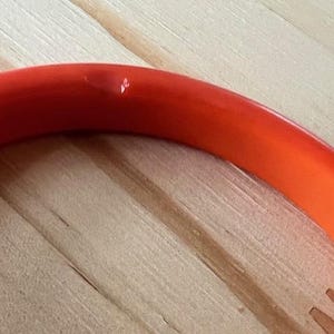 May include: A smooth, solid orange bangle bracelet with a rounded profile. The surface of the orange plastic material features a small, singular indentation or mark near the top curve, showing a slight variation in the otherwise uniform color.