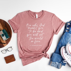 Puede incluir: Una camiseta rosa polvorienta con un gráfico blanco que dice "Be who God meant you to be and you will set the world on fire" y el texto "ST CATHERINE OF SIENA". La camiseta está colocada sobre una superficie blanca con un par de pantalones vaqueros azules, zapatillas blancas, una bandeja de madera con una planta y una cámara, un reloj, un bolígrafo y una cartera de cuero marrón.