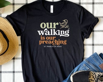 May include: Black t-shirt with a white, green, and orange graphic that says "Our walking is our preaching" with a small bird graphic and the text "St. Francis of Assisi".