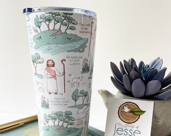 May include: A white tumbler with a blue and green design featuring a shepherd and sheep. The tumbler has the text "The Lord is my shepherd" and "He leads me to still waters" printed on it. The tumbler is sitting on a table with a wooden rosary and a book.