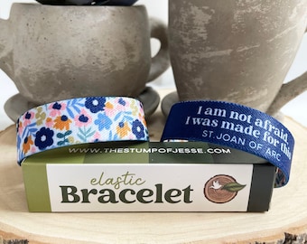 May include: Two elastic bracelets with floral and inspirational text designs rest on a wooden slice. One bracelet features a blue floral pattern, while the other is navy blue with the text "I am not afraid, I was made for this. ST. JOAN OF ARC."