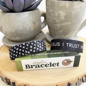 May include: Two black elastic bracelets with white designs. One bracelet has a white cross pattern and the other says "JESUS I TRUST IN". The bracelets are on a wooden surface with a small box that says "elastic Bracelet" and has a white dove with an olive branch on it.