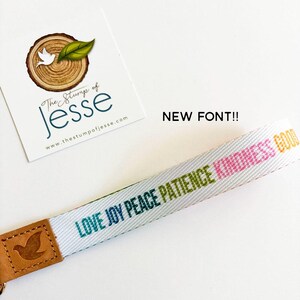 May include: A white fabric wristlet strap featuring the words LOVE, JOY, PEACE, PATIENCE, and KINDNESS printed in various colors. The strap is attached to a tan leather end piece embossed with a bird icon. A card with the text The Stump of Jesse is visible.