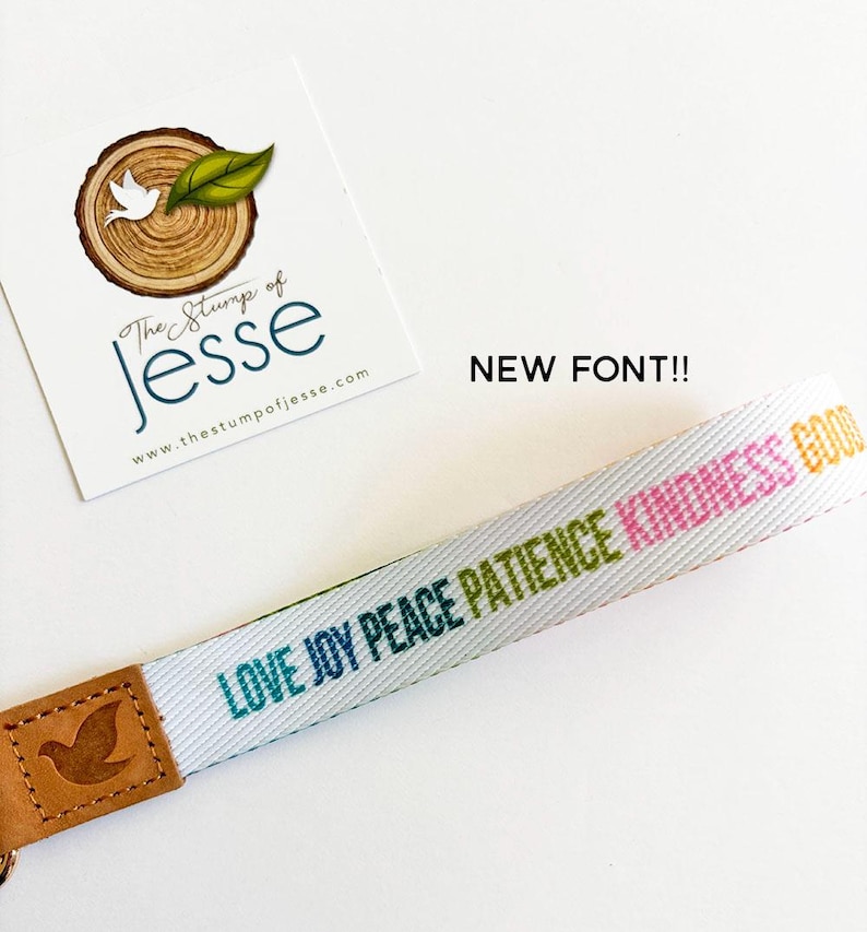May include: A white fabric wristlet strap featuring the words LOVE, JOY, PEACE, PATIENCE, and KINDNESS printed in various colors. The strap is attached to a tan leather end piece embossed with a bird icon. A card with the text The Stump of Jesse is visible.