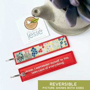 Puede incluir: Un llavero rectangular de tela con una anilla de metal plateado. Un lado presenta un colorido estampado floral con flores rojas, azules y amarillas sobre un fondo claro. El otro lado es rojo con el texto en blanco: Jesus, I surrender myself to You, take care of everything.