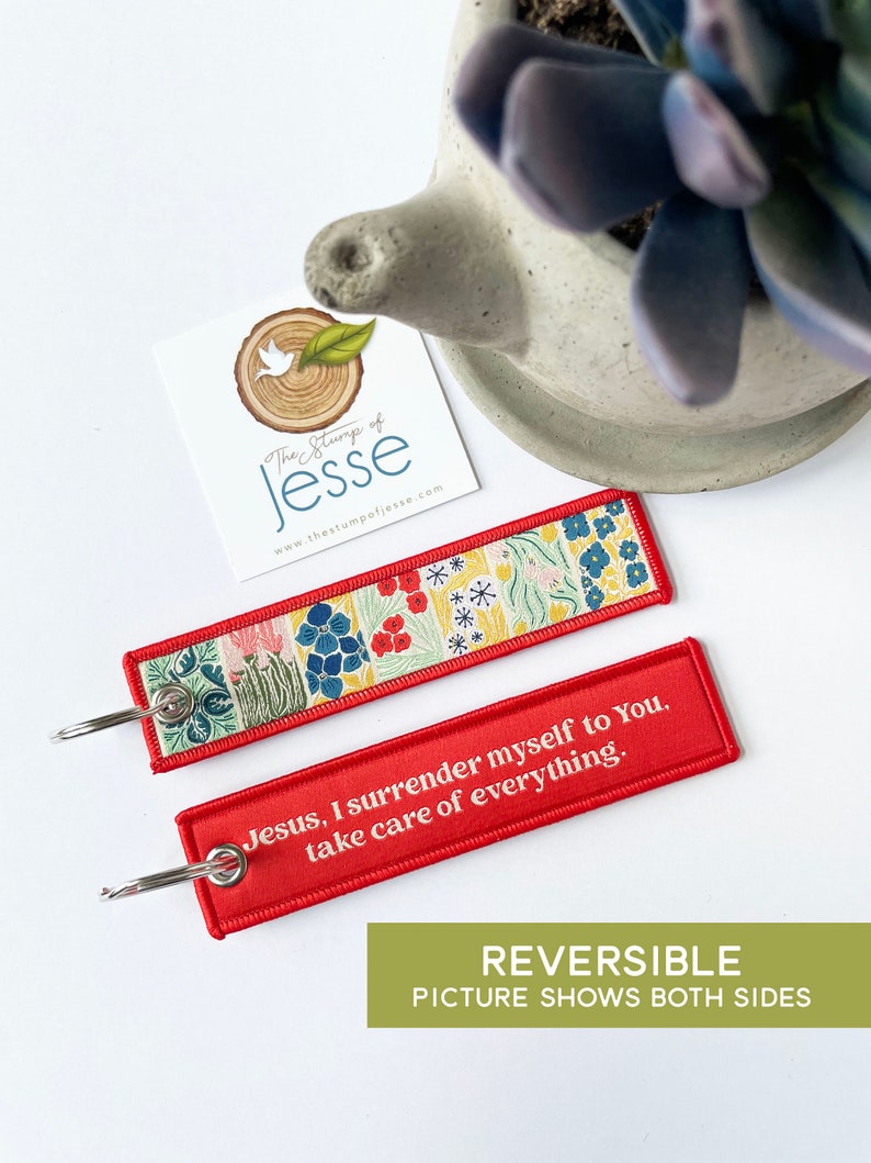 Puede incluir: Un llavero rectangular de tela con una anilla de metal plateado. Un lado presenta un colorido estampado floral con flores rojas, azules y amarillas sobre un fondo claro. El otro lado es rojo con el texto en blanco: Jesus, I surrender myself to You, take care of everything.