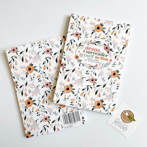 May include: A white journal with a floral pattern and the text "Jesus I surrender myself to you take care of everything". The journal is closed and lying on a white surface.