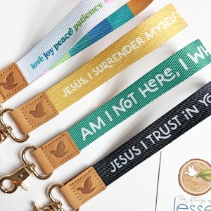 May include: Four fabric wristlet keychains with gold-tone metal lobster clasps and tan leather tabs embossed with a dove. The straps feature text including "love joy peace patience," "JESUS, I SURRENDER MYSELF," "AM I NOT HERE, I WILL," and "JESUS I TRUST IN YOU."