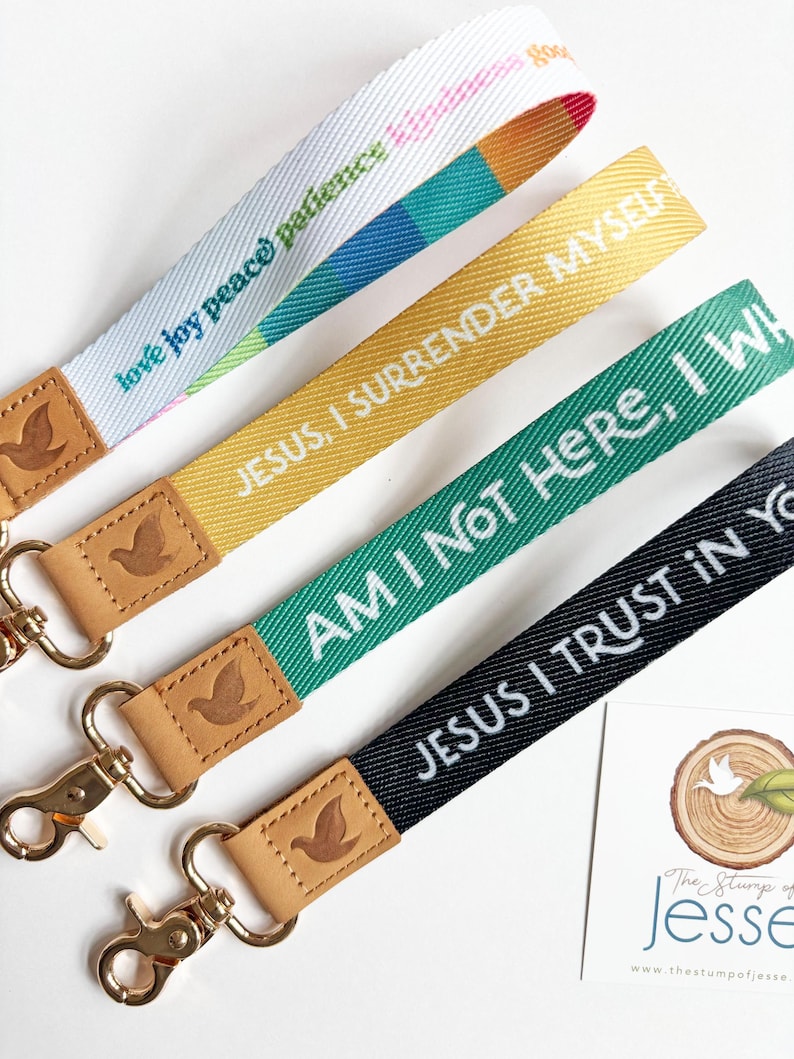 May include: Four fabric wristlet keychains with gold-tone metal lobster clasps and tan leather tabs embossed with a dove. The straps feature text including "love joy peace patience," "JESUS, I SURRENDER MYSELF," "AM I NOT HERE, I WILL," and "JESUS I TRUST IN YOU."