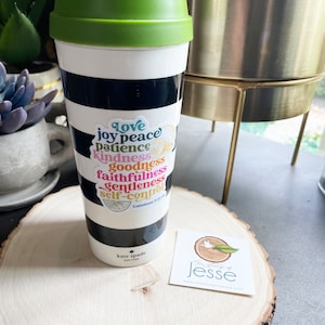 May include: A white and black striped travel mug with a green lid. A sticker with the words "love, joy, peace, patience, kindness, goodness, faithfulness, gentleness, self-control" is on the mug. The sticker also includes a lemon and a bible verse reference. The mug is sitting on a wooden surface with a small business card.
