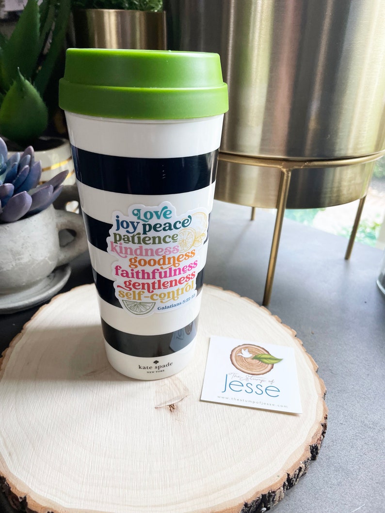May include: A white and black striped travel mug with a green lid. A sticker with the words "love, joy, peace, patience, kindness, goodness, faithfulness, gentleness, self-control" is on the mug. The sticker also includes a lemon and a bible verse reference. The mug is sitting on a wooden surface with a small business card.