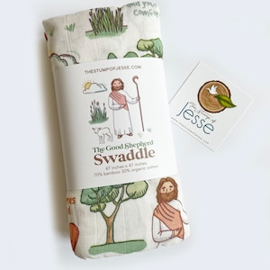 May include: A white swaddle blanket with a green, brown, and orange illustration of a shepherd with a staff and a lamb. The text "The Good Shepherd Swaddle" is printed on the blanket, along with the size and fabric composition: "47 inches x 47 inches, 70% bamboo 30% organic cotton".