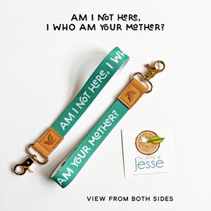 May include: Two green fabric wristlet keychains with tan leather accents and gold-tone metal swivel clasps. The fabric features the white text AM I NOT HERE, I WHO AM YOUR MOTHER? A square white card with a tree stump logo and text is included.