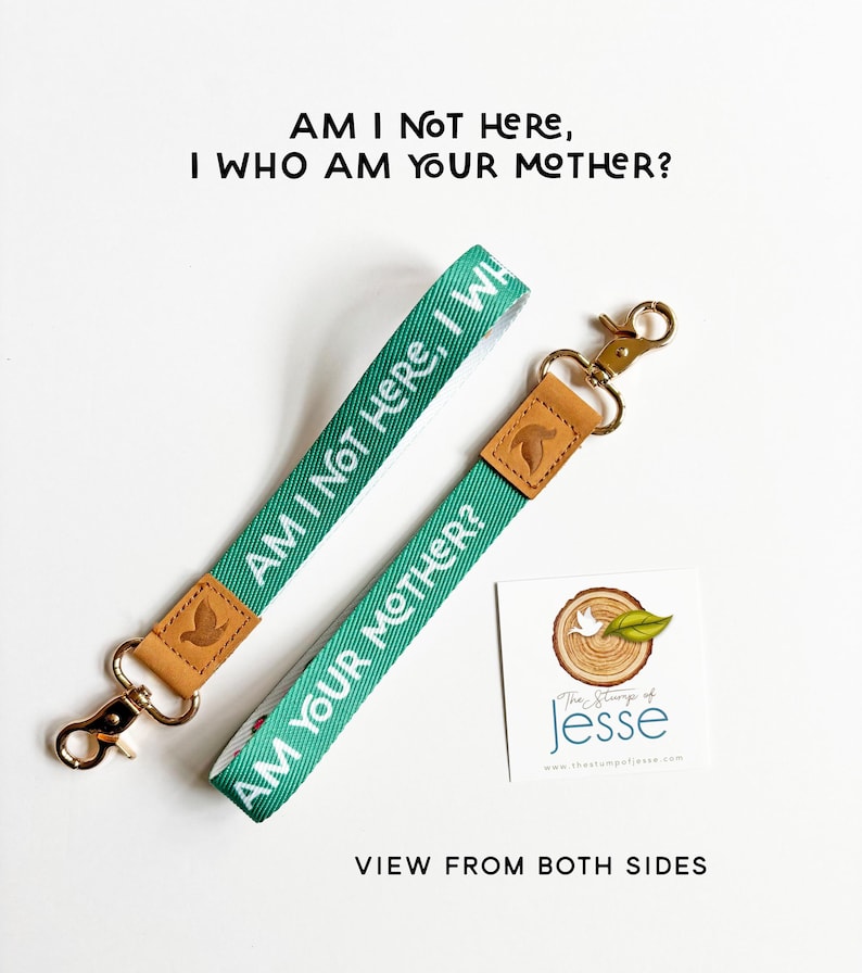 May include: Two green fabric wristlet keychains with tan leather accents and gold-tone metal swivel clasps. The fabric features the white text AM I NOT HERE, I WHO AM YOUR MOTHER? A square white card with a tree stump logo and text is included.