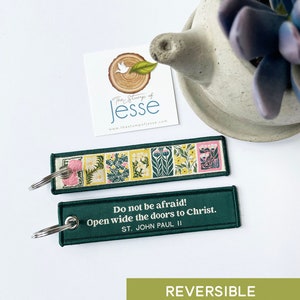 Puede incluir: Un llavero rectangular de tela con una anilla de metal. Un lado presenta una serie de paneles florales coloridos en tonos verde, amarillo y rosa. El otro lado es verde oscuro con el texto en blanco: Do not be afraid! Open wide the doors to Christ. ST. JOHN PAUL II.