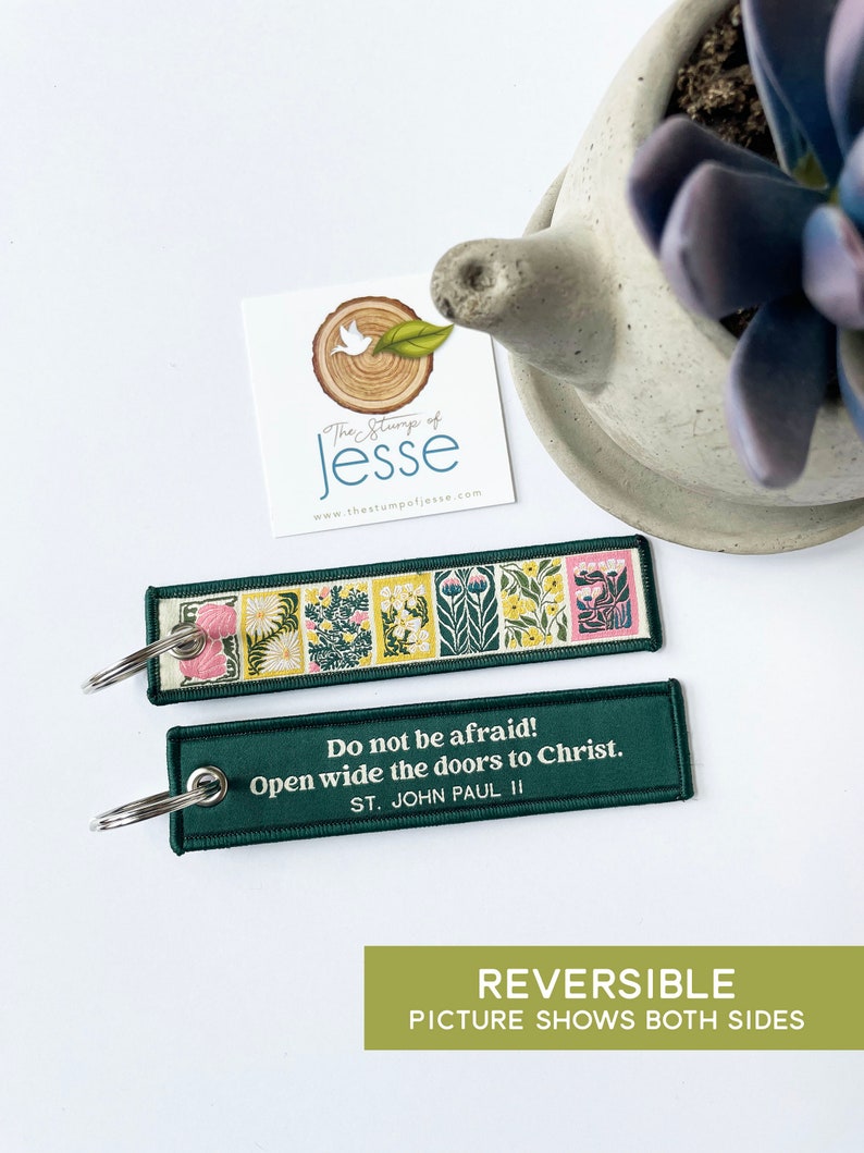 Puede incluir: Un llavero rectangular de tela con una anilla de metal. Un lado presenta una serie de paneles florales coloridos en tonos verde, amarillo y rosa. El otro lado es verde oscuro con el texto en blanco: Do not be afraid! Open wide the doors to Christ. ST. JOHN PAUL II.