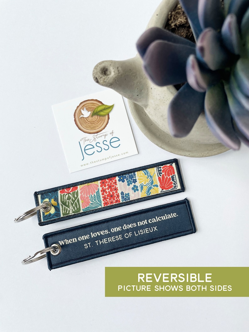 Puede incluir: Dos llaveros rectangulares de tela con anillas de metal. Un lado presenta un colorido estampado floral con motivos en rojo, rosa, amarillo y verde. El otro lado es azul oscuro con el texto en blanco: When one loves, one does not calculate. ST. THERESE OF LISIEUX.