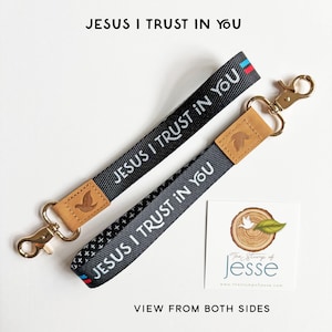 May include: Two black fabric wristlet keychains featuring the white text JESUS I TRUST IN YOU. Each strap has a tan leather accent embossed with a dove icon and a gold-tone metal swivel lobster clasp. One side shows a solid black weave, while the other displays a small white cross pattern with red and blue stripes at the end.