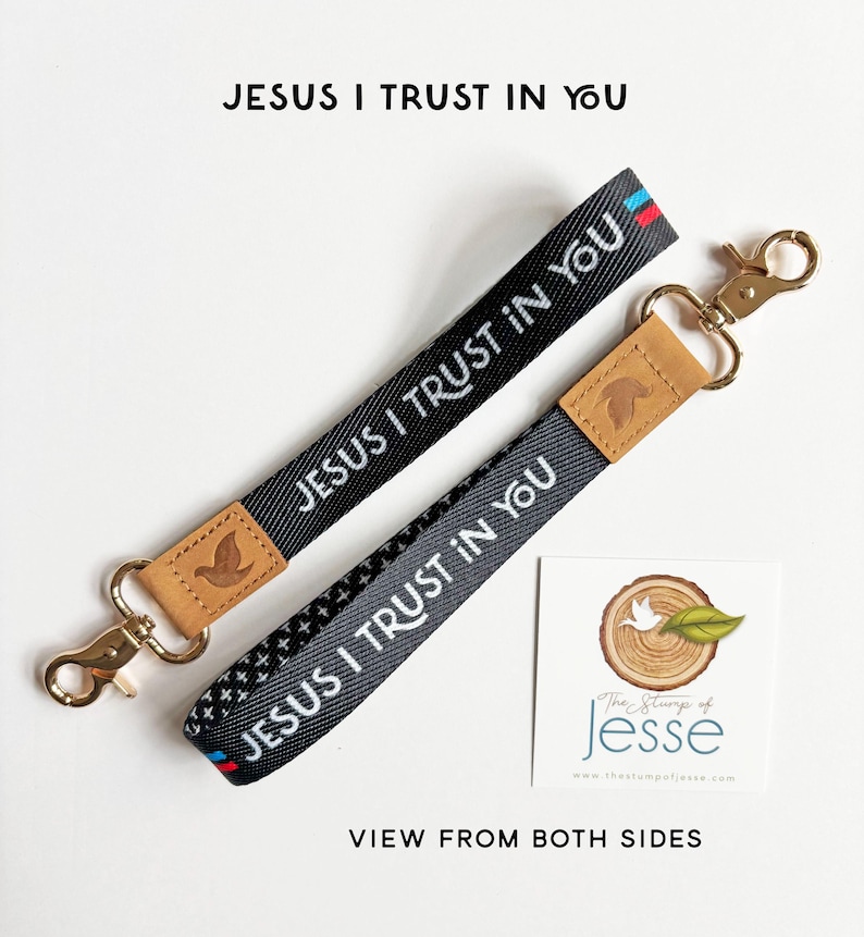 May include: Two black fabric wristlet keychains featuring the white text JESUS I TRUST IN YOU. Each strap has a tan leather accent embossed with a dove icon and a gold-tone metal swivel lobster clasp. One side shows a solid black weave, while the other displays a small white cross pattern with red and blue stripes at the end.