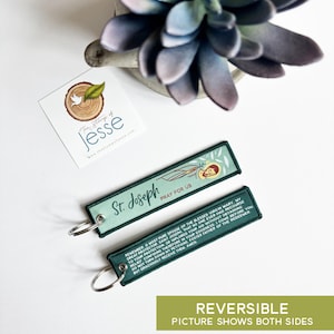 Puede incluir: Dos llaveros rectangulares de tela con bordes cosidos en verde oscuro y anillas de metal plateado. Un lado presenta una ilustraci&oacute;n de San Jos&eacute; con el texto St. Joseph PRAY FOR US. El reverso muestra una oraci&oacute;n en texto blanco.