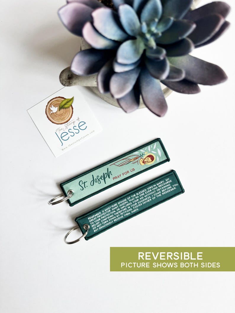 Puede incluir: Dos llaveros rectangulares de tela con bordes cosidos en verde oscuro y anillas de metal plateado. Un lado presenta una ilustraci&oacute;n de San Jos&eacute; con el texto St. Joseph PRAY FOR US. El reverso muestra una oraci&oacute;n en texto blanco.