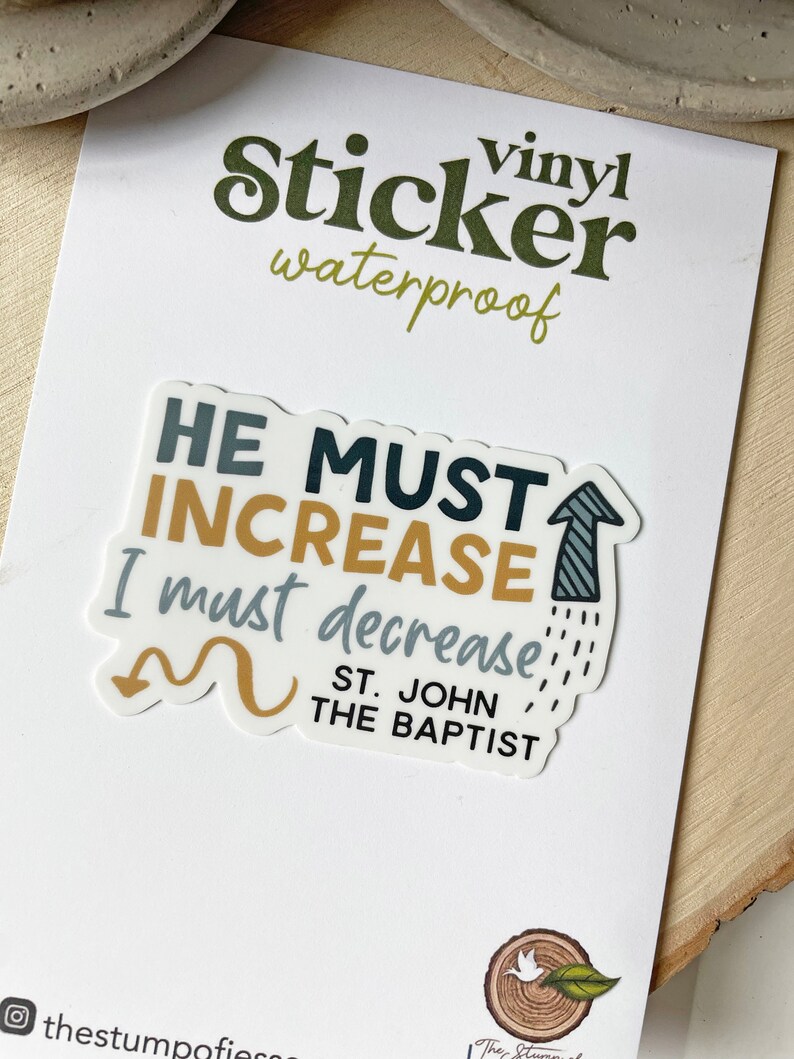 May include: A vinyl sticker with a white background and a blue, gold, and black design. The sticker features the text "HE MUST INCREASE I must decrease" with a gold arrow pointing up and a gold arrow pointing down. The text "ST. JOHN THE BAPTIST" is also included in the design.
