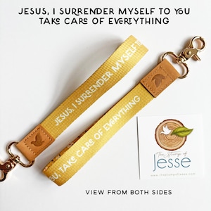 May include: A yellow fabric wristlet keychain featuring the white text "JESUS, I SURRENDER MYSELF TO YOU" and "TAKE CARE OF EVERYTHING". It includes a tan leather accent piece embossed with a dove and a gold-toned metal lobster clasp.