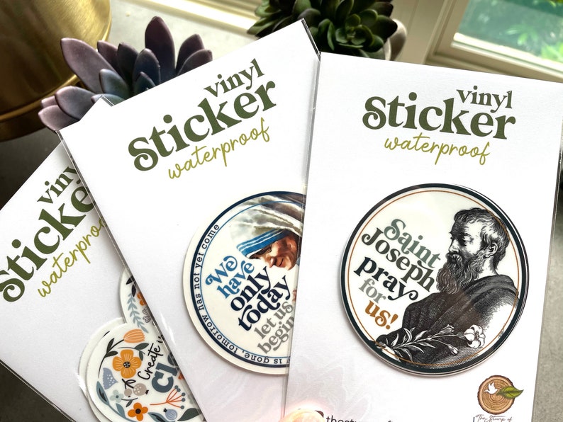 May include: Three white cards with the text "vinyl sticker waterproof" printed in green. Each card features a round sticker with a different design. The first sticker features a floral design with the text "Create a life you love". The second sticker features a portrait of Mother Teresa with the text "We have only today, let us begin". The third sticker features a portrait of Saint Joseph with the text "Saint Joseph pray for us!"
