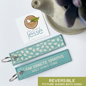 Puede incluir: Dos llaveros rectangulares de tela en azul claro con una anilla de metal plateado. Un lado presenta un patr&oacute;n repetitivo de peque&ntilde;os p&aacute;jaros blancos. El otro lado muestra el texto blanco "VENI SANCTE SPIRITUS" y "HOLY SPIRIT COME" en letra clara.
