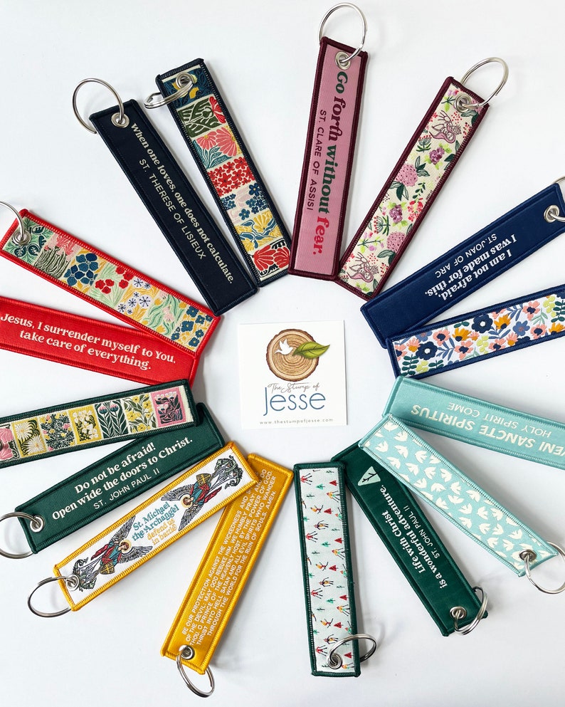 Puede incluir: Una colecci&oacute;n de llaveros de tela rectangulares dispuestos en c&iacute;rculo, con varios colores, patrones florales y citas religiosas. Cada llavero incluye una anilla de metal. Una tarjeta cuadrada en el centro muestra el texto The Stump of Jesse.