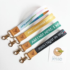 May include: Four fabric wristlet keychains with gold-tone metal swivel clasps and tan leather accents embossed with a dove. The straps feature text including love, joy, peace, patience, kindness, Jesus, I surrender myself, Am I not here, I who, and Jesus I trust in you.