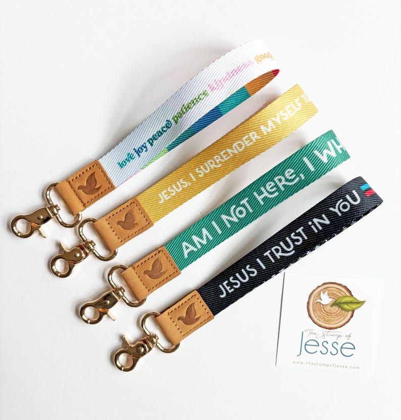 May include: Four fabric wristlet keychains with gold-tone metal swivel clasps and tan leather accents embossed with a dove. The straps feature text including love, joy, peace, patience, kindness, Jesus, I surrender myself, Am I not here, I who, and Jesus I trust in you.