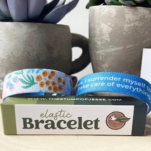 May include: Two elastic bracelets, one with a floral pattern and the other with the text "us, I surrender myself to take care of everything."  The bracelets are on a wooden surface with a box that says "elastic Bracelet" and the website address "www.thestumpofjesse.com".