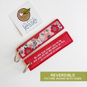 Puede incluir: Dos llaveros rectangulares con anillas de metal plateado. Un lado presenta un estampado floral con flores rojas, rosas y verdes sobre fondo blanco. El otro lado es de color rojo s&oacute;lido con texto en blanco: Be who God meant you to be and you will set the world on fire, ST. CATHERINE OF SIENA.