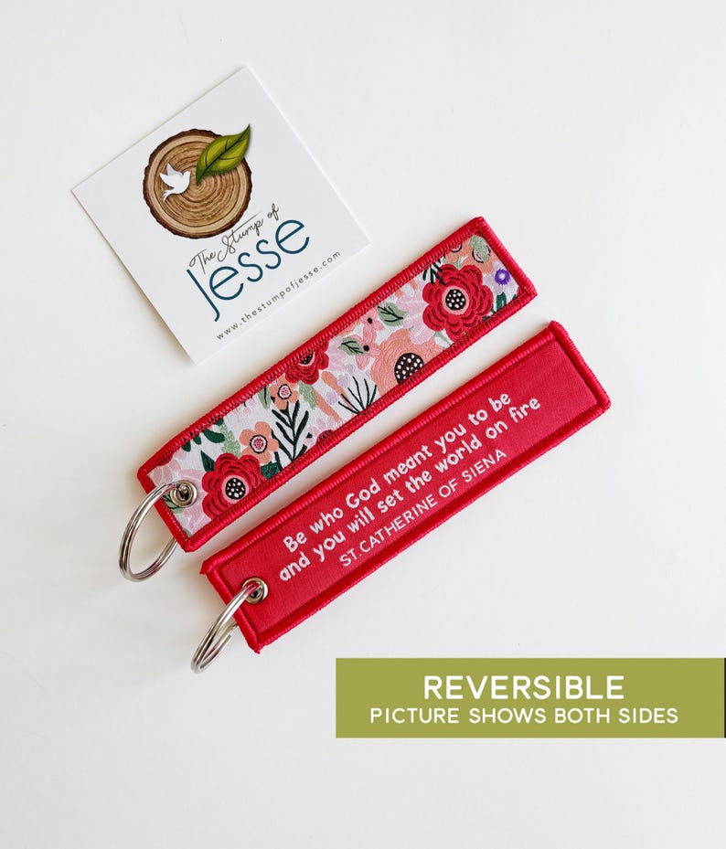 Puede incluir: Dos llaveros rectangulares con anillas de metal plateado. Un lado presenta un estampado floral con flores rojas, rosas y verdes sobre fondo blanco. El otro lado es de color rojo s&oacute;lido con texto en blanco: Be who God meant you to be and you will set the world on fire, ST. CATHERINE OF SIENA.