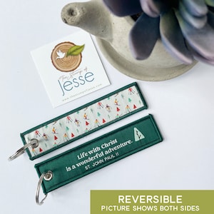 Puede incluir: Un llavero rectangular de tela con una anilla de metal. Un lado presenta un fondo blanco con un patr&oacute;n colorido de esquiadores y &aacute;rboles. El otro lado es verde oscuro con el texto en blanco: Life with Christ is a wonderful adventure. ST. JOHN PAUL II.