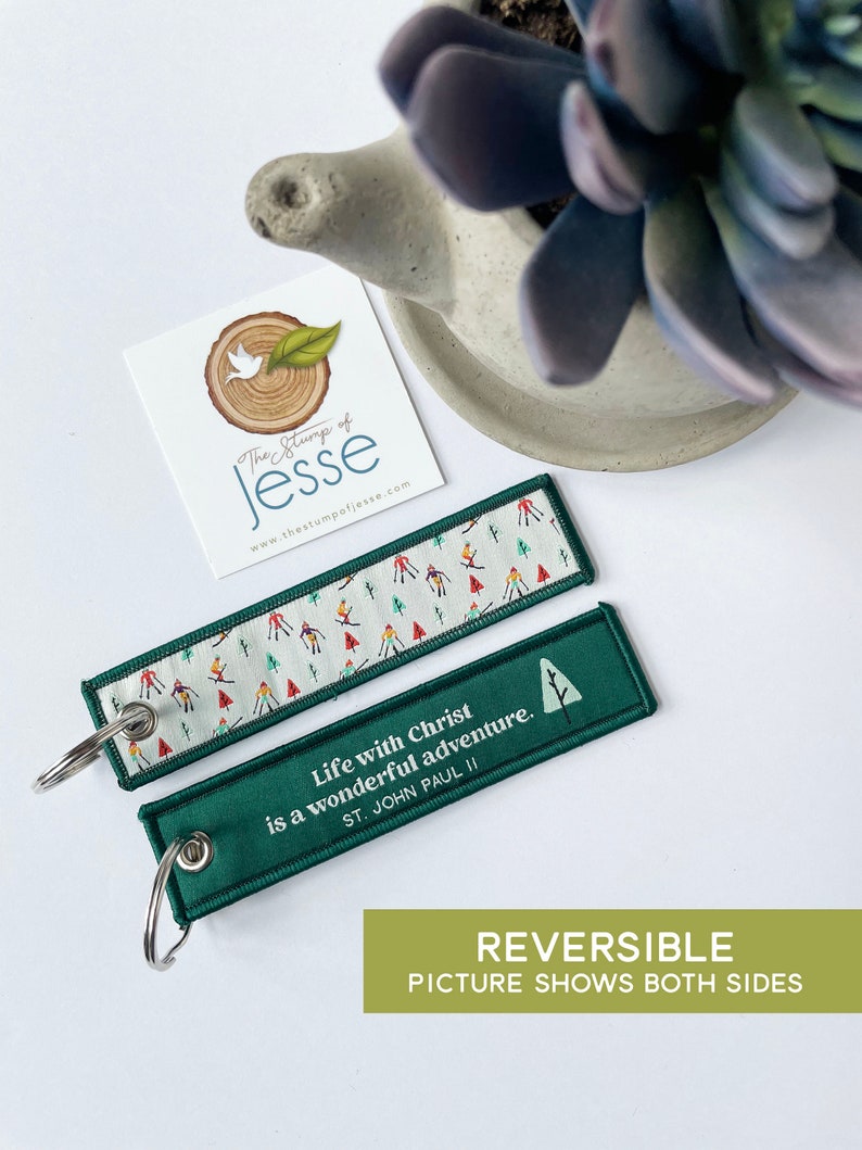 Puede incluir: Un llavero rectangular de tela con una anilla de metal. Un lado presenta un fondo blanco con un patr&oacute;n colorido de esquiadores y &aacute;rboles. El otro lado es verde oscuro con el texto en blanco: Life with Christ is a wonderful adventure. ST. JOHN PAUL II.