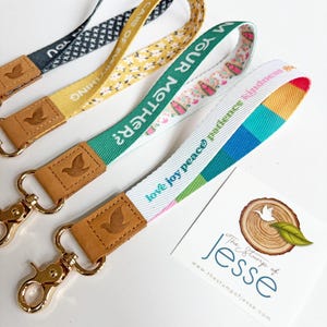 May include: Four fabric wristlet lanyards with gold-tone metal lobster clasps and tan leather tabs embossed with a bird icon. The straps feature various patterns and text including "take care of everything," "am your mother?" and "love joy peace."