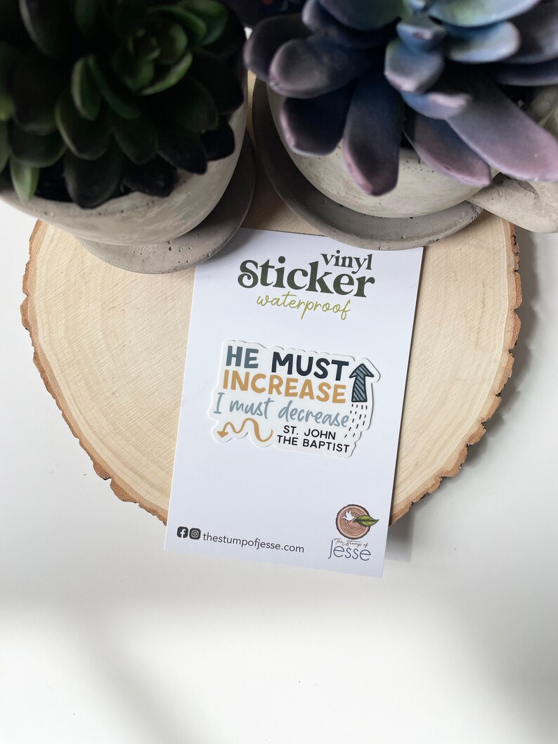 May include: A vinyl sticker with the text "HE MUST INCREASE I must decrease" and "ST. JOHN THE BAPTIST" in a colourful, handwritten style. The sticker is on a wooden surface.
